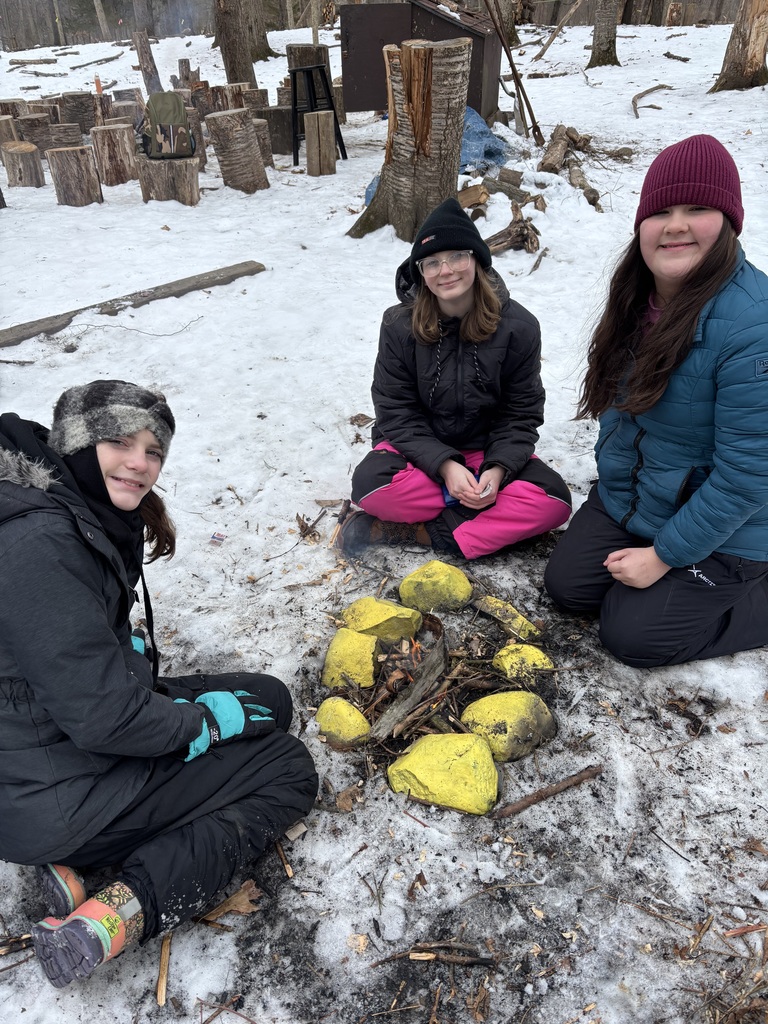Three sixth grade girls sit around the fire that they built. The fire is in a small fire pit that is surrounded by large rocks that have been painted yellow. There is still snow on the ground.