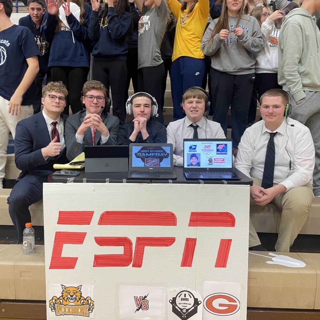 Five students are sitting on the bleachers with mock headsets on. They have a homemade sign in front of them that says ESPN and has the Woodmore and Gibsonburg logos on it. All of the students are in business casual attire to appear as on air sports analysts.