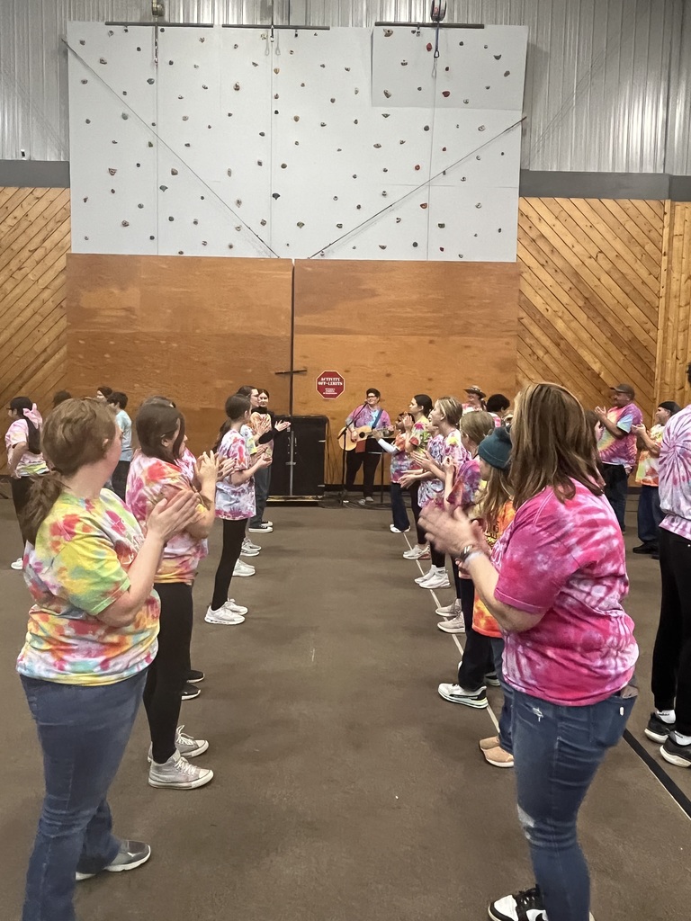 Students are in two lines facing each other in the gymnasium for square dancing. There is a band at the far end of the gym playing the music for the dance.