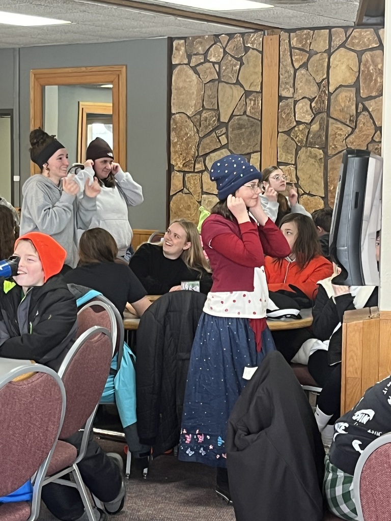 Students are in the dining hall at circular tables playing games before their meal begins. Four people are standing holding on to their ears.