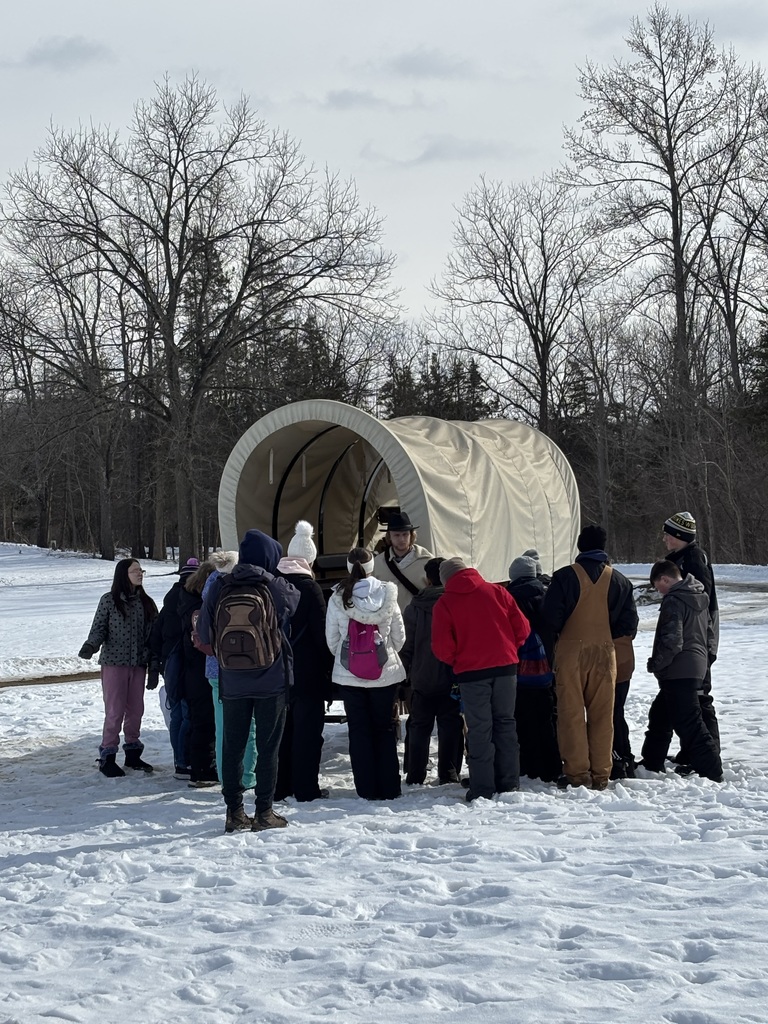 A group of students stands in front of a reenactor, looking at a covered wagon in the snow.