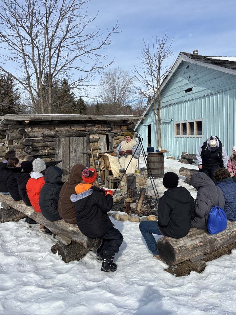 Sixth grade students are sitting on logs in front of a log cabin with a fire outside and a reenactor speaking to the students by the fire.