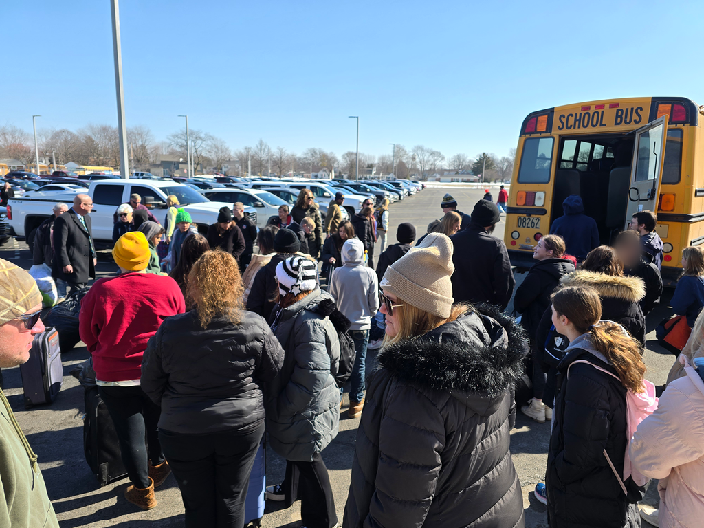 Students and parents are grouped in the parking lot around the back of the luggage bus, waiting for their student's luggage to be offloaded.