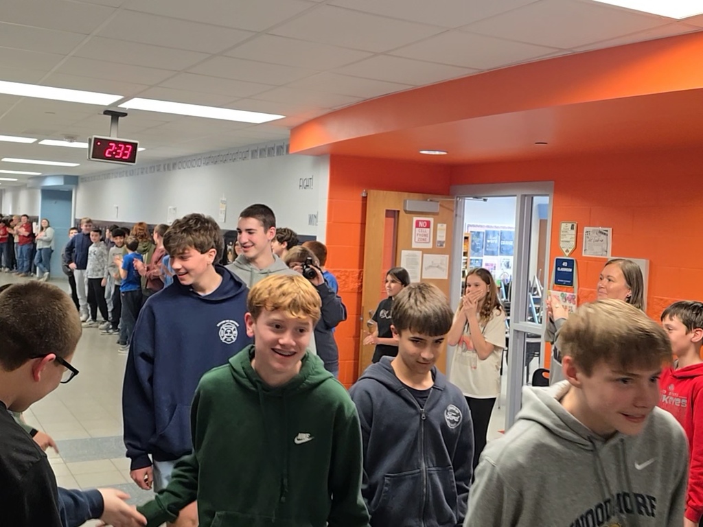 Boys 8th grade basketball team walks down the hallway lined with students and teachers who are clapping. The athletes give high fives as they walk past.