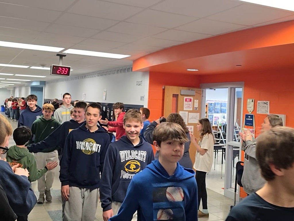 Boys 8th grade basketball team walks down the hallway lined with students and teachers who are clapping. The athletes give high fives as they walk past.