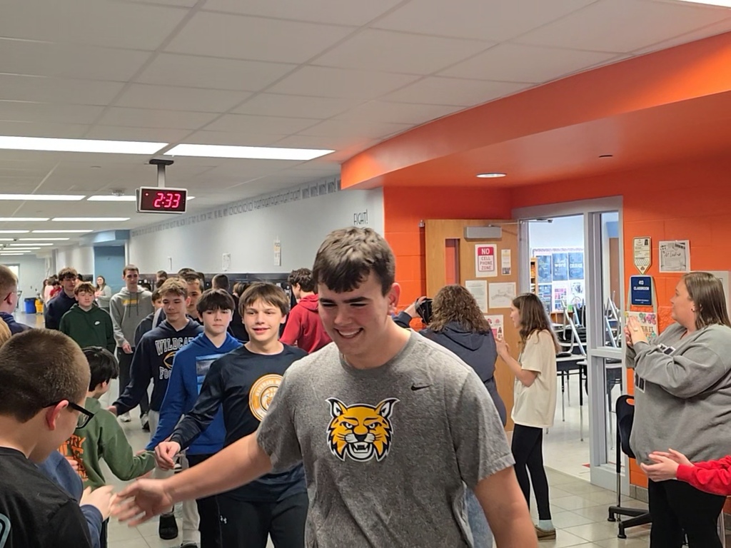 Boys 8th grade basketball team walks down the hallway lined with students and teachers who are clapping. The athletes give high fives as they walk past.