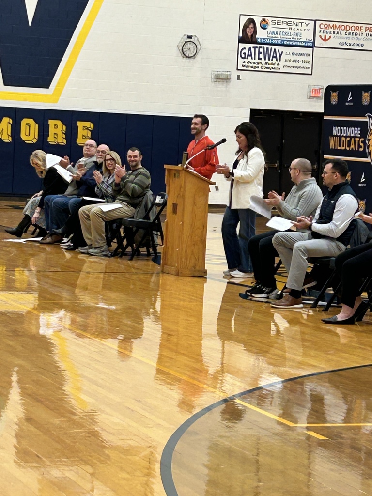 A woman is standing at the podium in the high school gym speaking, while Mr. Avers stands next to her. Other participants in the assembly are seated in chairs on either side.