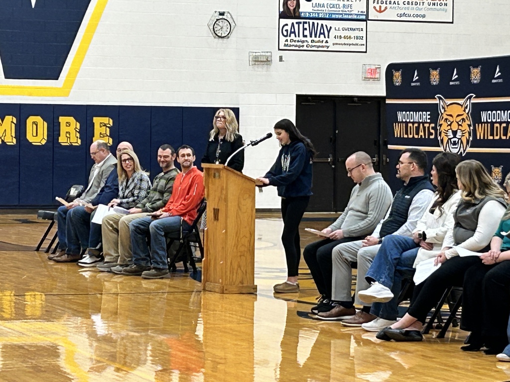 A student reads their nomination while standing at a podium in the gym. Other people sit on either side listening.