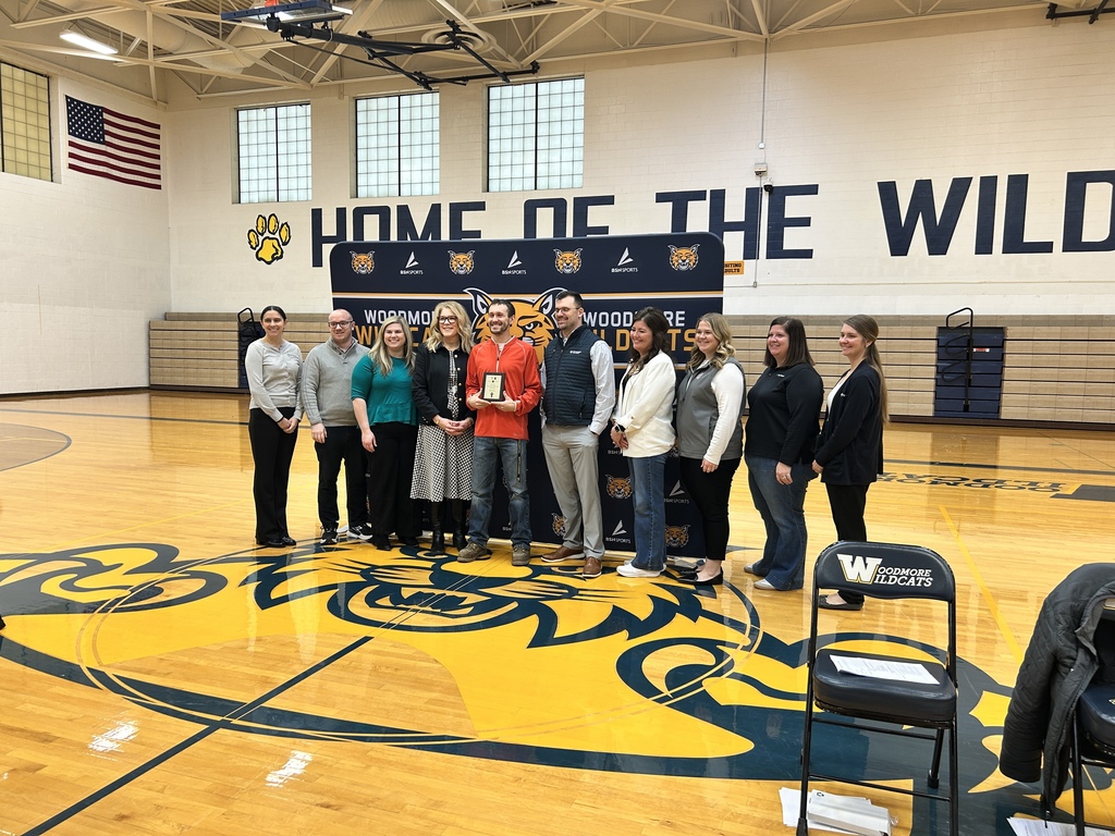 Mr. Avers stands in the gym in front of the Woodmore backdrop with the various representatives of the sponsoring companies that organize the Golden Owl Award and Mrs. Coil, the high school principal.