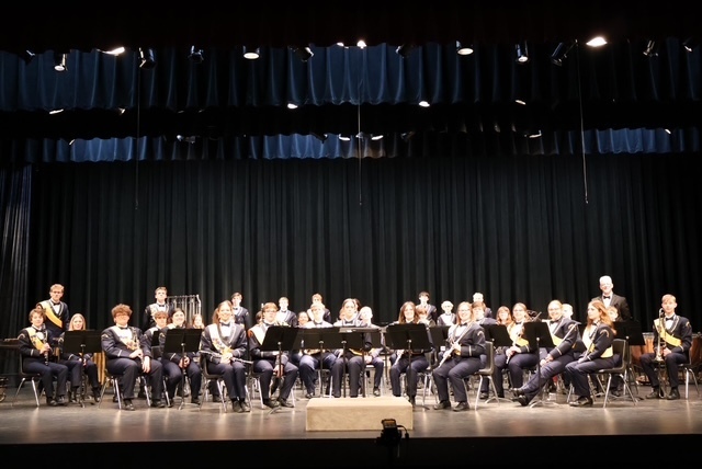 The high school band sits on the performance stage with their instruments and music stands. Mr. Emerine is in the back right.