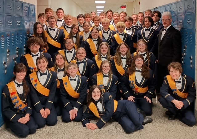 The high school band members pose in a hallway. Students in the front are kneeling or laying on the floor, while students in the back are standing. Mr. Emerine is on the right. All students are in their concert band uniforms (blue suits with gold trim, white shirts, black bow tie, and gold sash). 