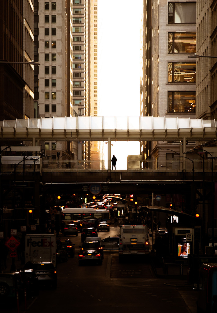 Photo by Logan Campbell looking down a street in a busy urban area. Light is coming between the skyscrapers, illuminating a person standing on a pedestrian walkway that is going over the traffic on the street.