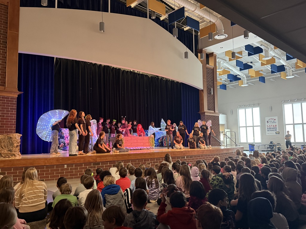 The Little Mermaid cast performing on stage. The cast is looking at our Ariel and Eric in a boat in the center of the stage. There are students with  lit up wings surrounding them in the back.