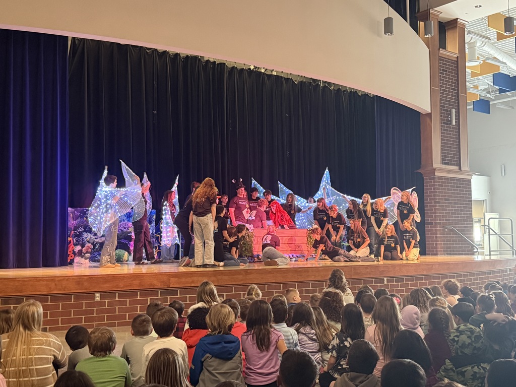 The Little Mermaid cast performing on stage. The cast is looking at our Ariel and Eric in a boat in the center of the stage. There are students with  lit up wings surrounding them in the back.