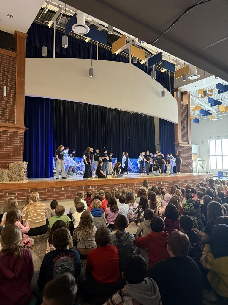The Little Mermaid cast on stage, surrounded by cast members wearing lit up wings. Elementary and Middle School students sit on the floor, watching the performance.