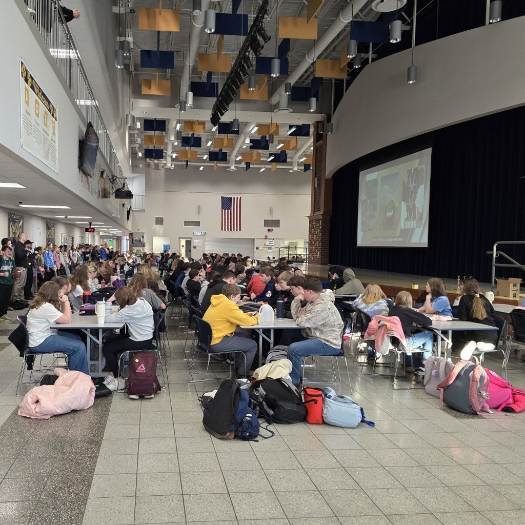 Middle School students sit in the cafetorium and stand along the back wall to watch the video on the pull down screen on the stage.