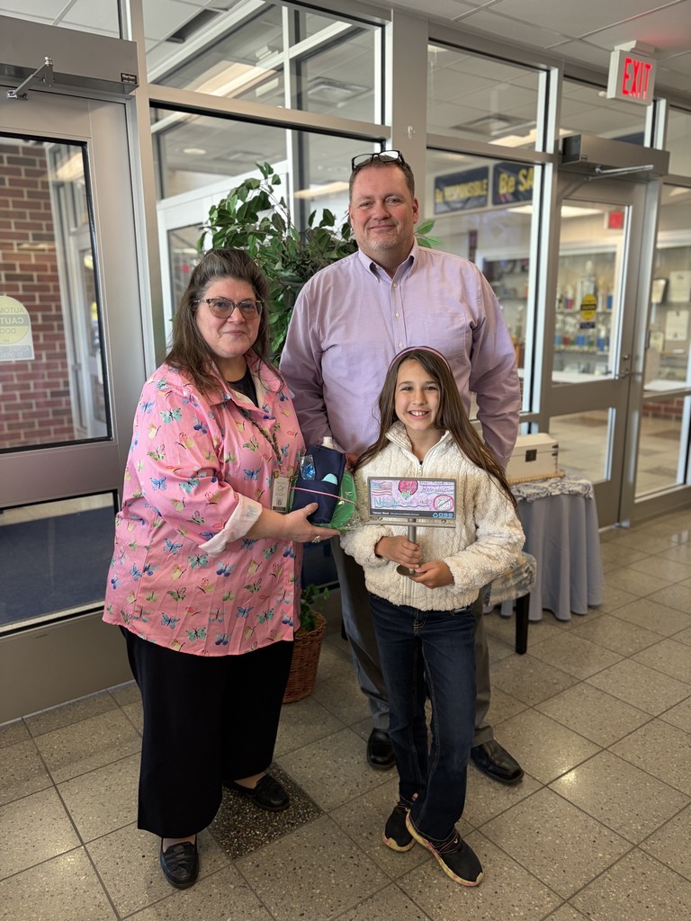Harper standing in the main office of the middle school receiving her mini billboard of her design along with other prizes from an OSS representative who is in a pink shirt on the left of the picture. Mr. Barrett stands behind Harper.