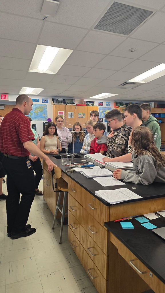 Students are gathered around the teacher's lab table at the front of the classroom while the teacher stands behind it, explaining procedure. There is a Bunsen burner on the table.