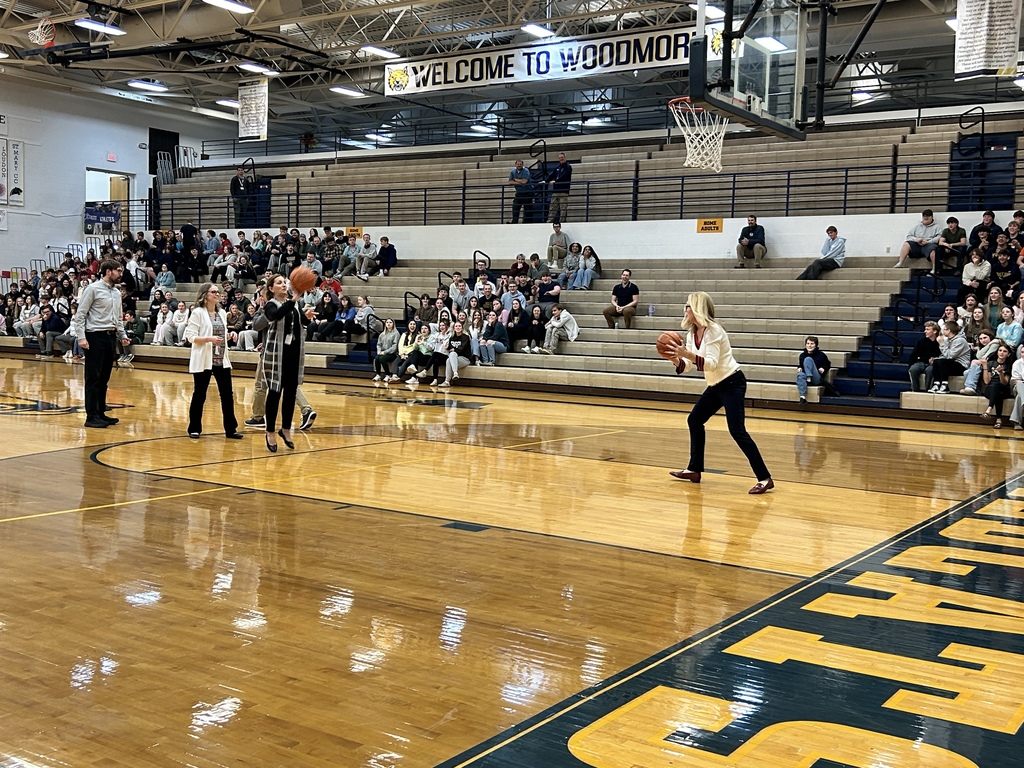 Students are in the background sitting on bleachers watching. A line of teachers is at the foul line waiting to shoot the basketball. Mrs. Coil has a basketball and is heading back to the foul line.
