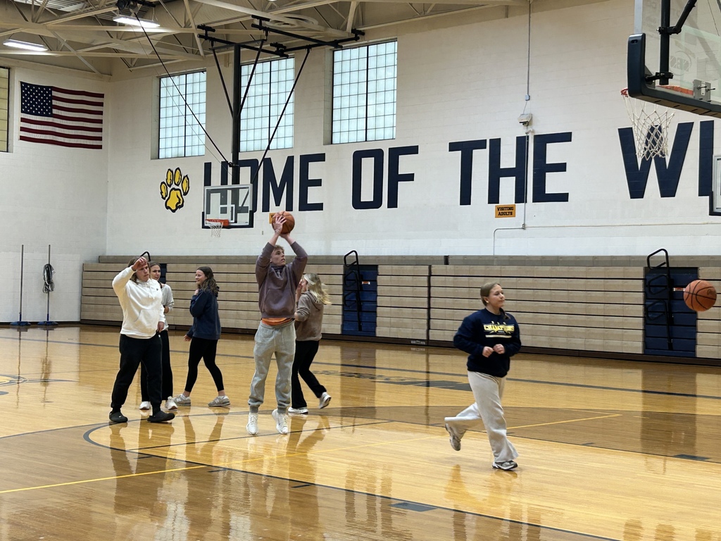 A student on the right is running to retrieve their basketball.  The student in the middle is in the process of jumping off the ground for a jump shot from the foul liine. Three more students wait for their turn.