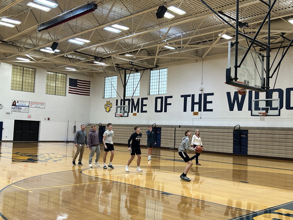 A line of students waiting for their turn to shoot a basketball are lined up at the foul line. Mrs. Coil is in the back watching. Mr. Black is at the end of the line.