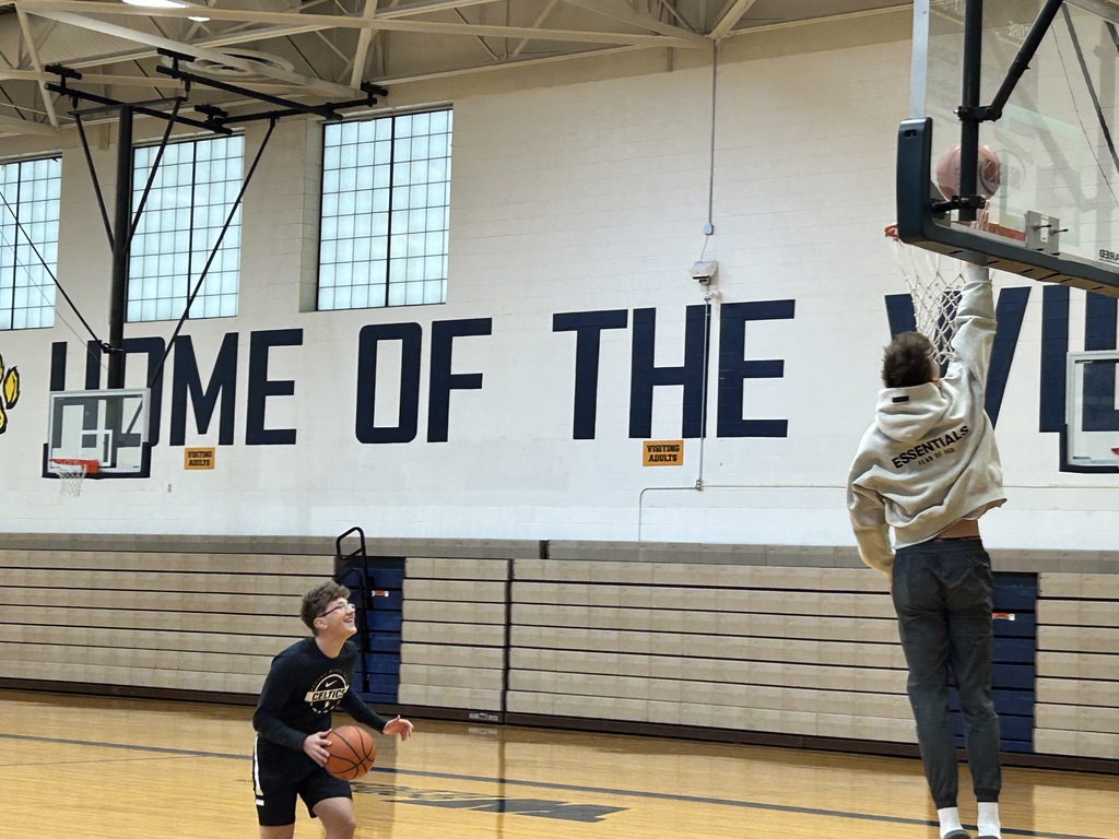 A student is seen jumping up for a layup. Another student is on the ground dribbling the ball, smiling toward the basket.