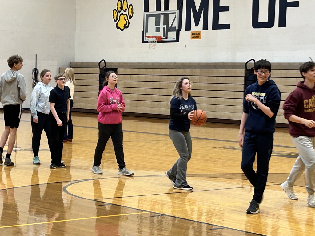 A close up of a line of students waiting to shoot basketballs. The two students on the right have already shot, the one in the middle has a ball and is getting ready and the one on the far left is waiting. You can see the empty Visitor's side bleachers in the back.