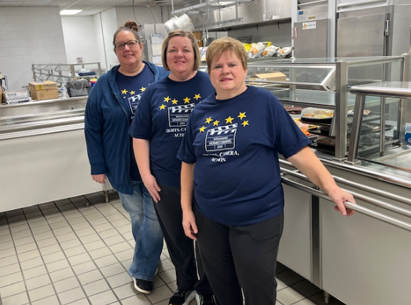 The high school lunch staff stand in the serving line to pose for a picture. The kitchen is behind them.