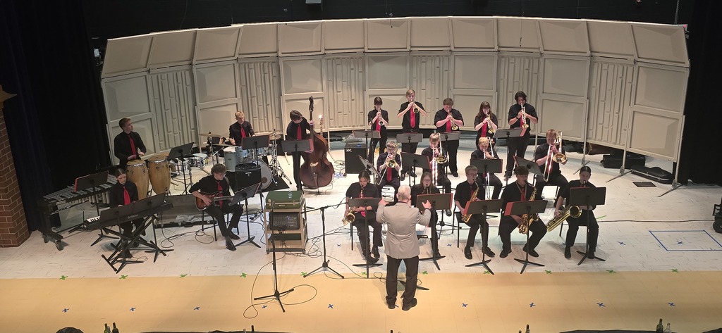 The jazz band is on the stage at the elementary school, playing their instruments with the band director standing at the front of the stage, conducting.
