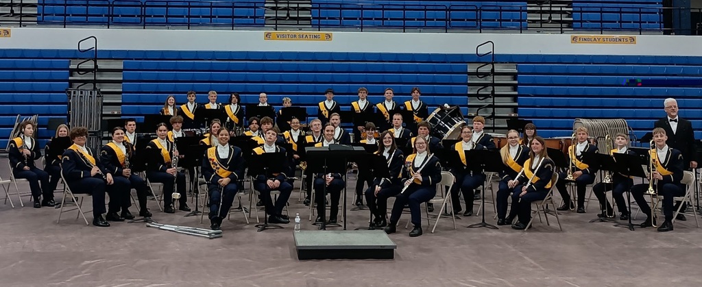Woodmore Concert band sitting in a gym, in concert formation with blue bleachers behind them. All are looking at the camera and smiling.
