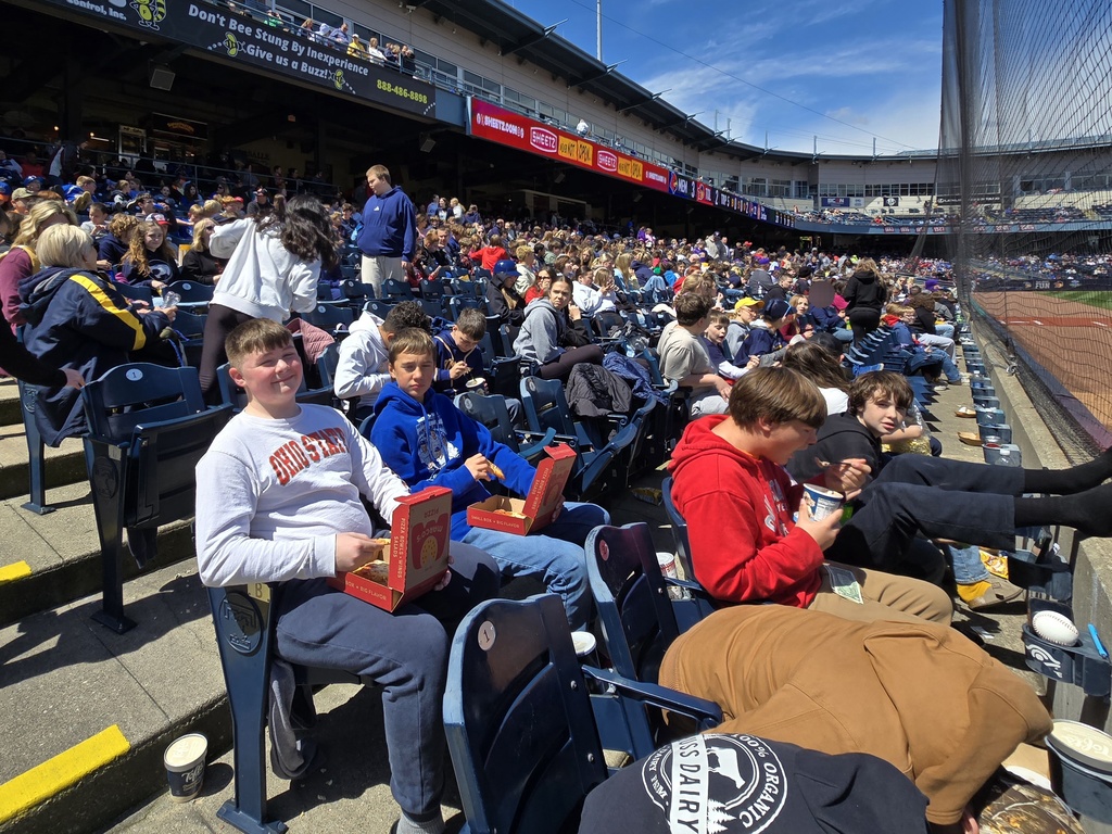 Students sit in the stadium seats watching the game. Some are eating pizza, some are eating ice cream, some are hiding from the camera. Blue skies and great weather!