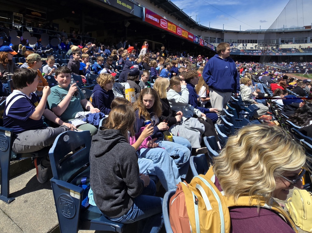 Students sit in the stadium seats watching the game. Some are drinking pop, some are eating ice cream, some are hiding from the camera. Blue skies and great weather!