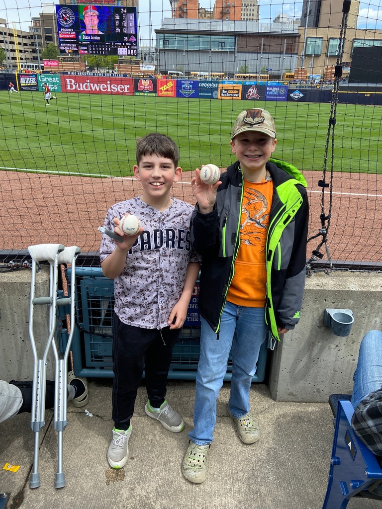 Nolan and Rhett posing with their foul balls that they caught during the game. They are standing in front of the netting with the ball field behind them.