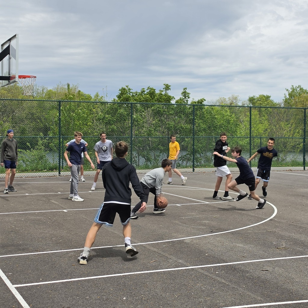 A group of students playing basketball on the courts at the park in Genoa. Trees and the river are in the background.