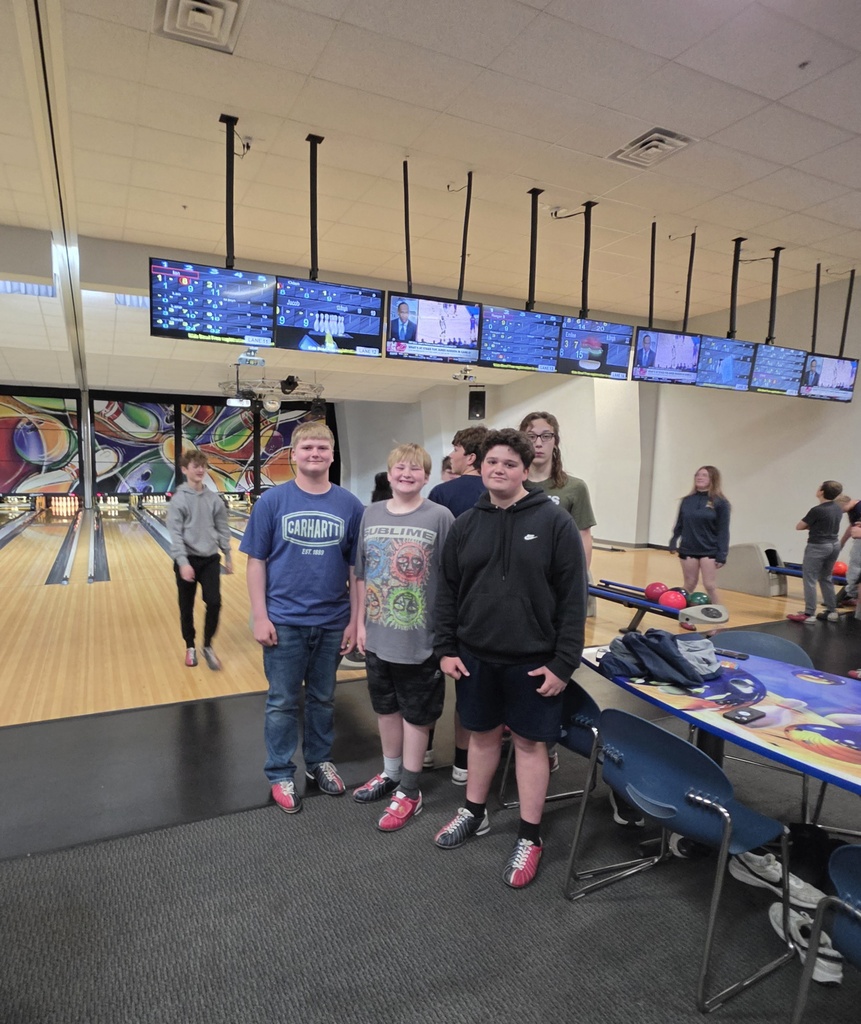 A group of students pose for a picture standing in front of the bowling lanes with the tables and TVs in the background.