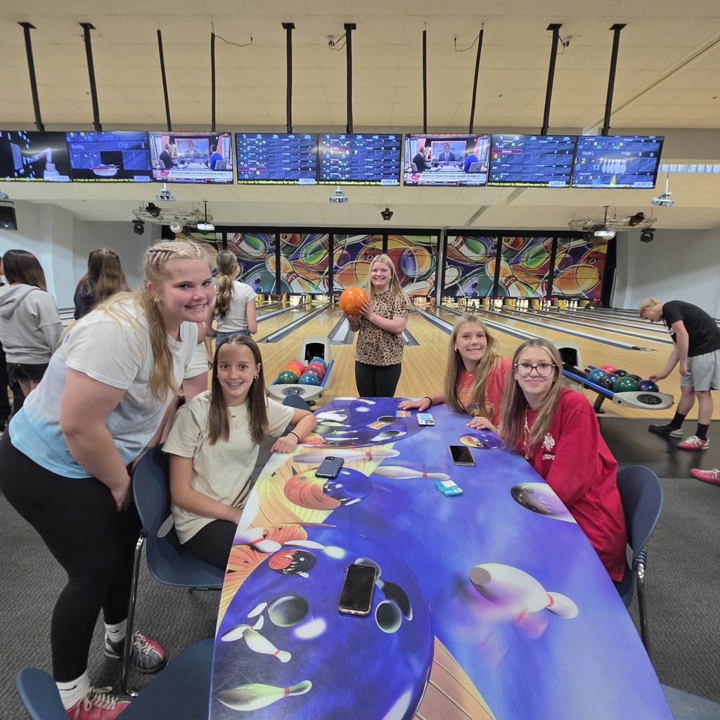 A group of students poses for a picture while seated at a table. The bowling alley and TVs are in the background.