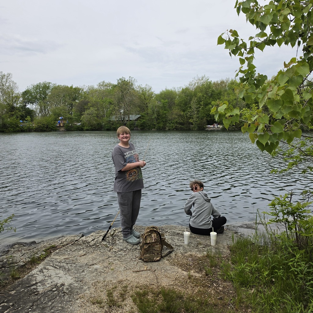 Two students turn to the camera while on the bank of the river. Both are fishing.