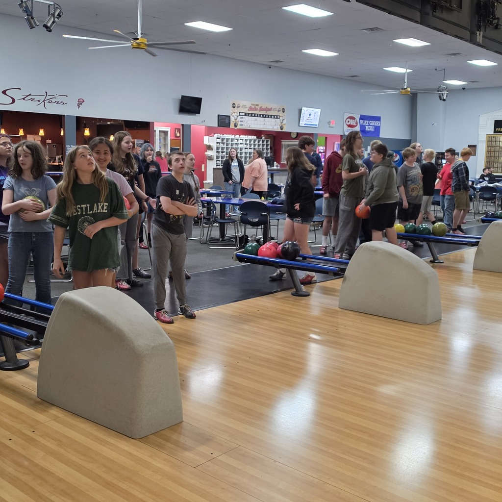 Standing on the wooden portion of the bowling alley, facing the students that are preparing to or waiting to bowl.