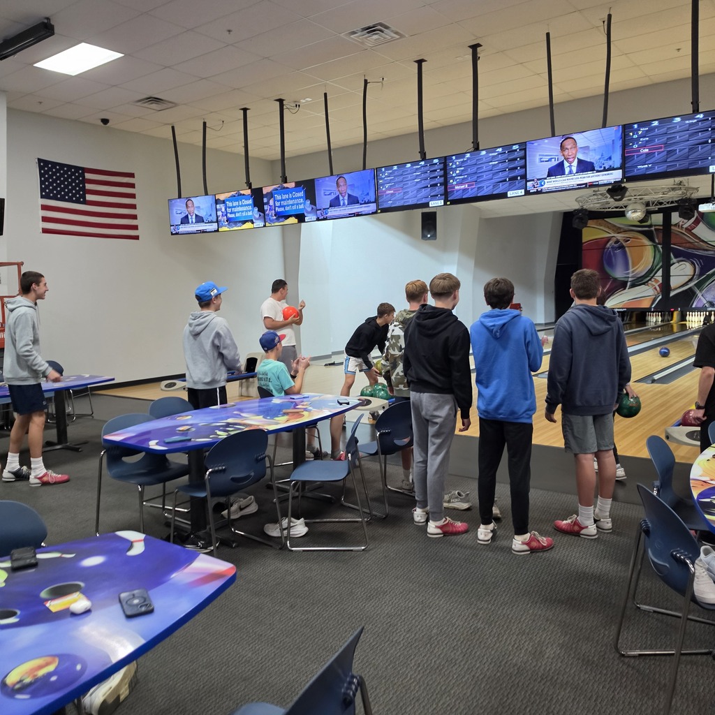 A bowling alley with students waiting on the carpet for their turn to bowl. TVs are overhead, some have the score, some are broadcasting programming.
