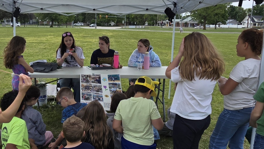 Three students sit at a table presenting about floriculture. A group of students sits and stands on the grass in front of them, listening and watching their demonstration.