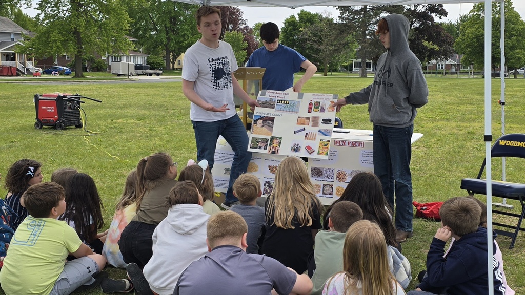 Two students are presenting about popcorn in front of elementary students seated on the grass. There is a third high school student in the background making popcorn.