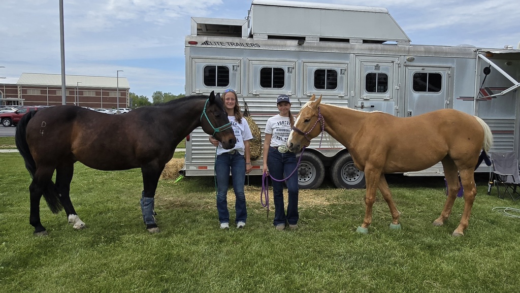 Two students stand in the center of the frame, holding the reins of two horses. On the left is a dark brown colored horse and on the right is a light tan colored horse. The trailer used to transport them is in the background.