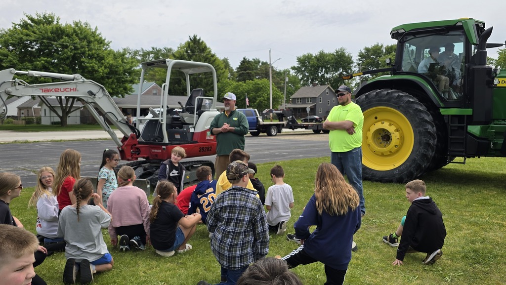 Two farmers stand in front of students with their heavy equipment in the background. A group of students sits on the grass in front of them, listening to their presentation.