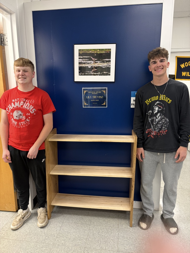 Two students stand on either side of a wooden shelving unit with three shelves.  They are standing against a dark blue wall in the high school office.