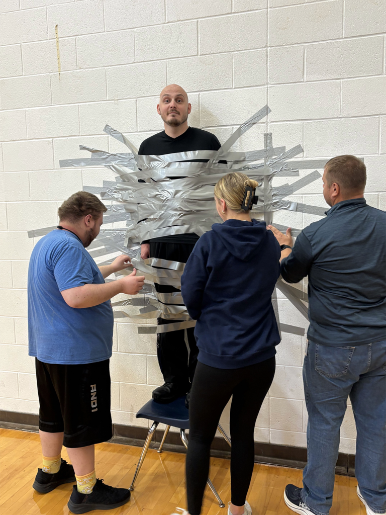 Office Psota is taped to a white wall in the gym with duct tape (his feet are standing on a chair). Two students and a teacher assist while Officer Psota has a silly grin on his face.