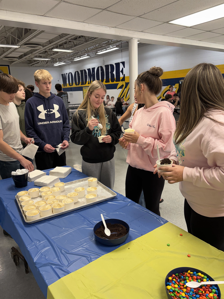 There is a table with blue and yellow tablecloths in the foreground of the picture with ice cream cups and sundae toppings on it. Students have made a line and are going through past the table making their own sundaes.