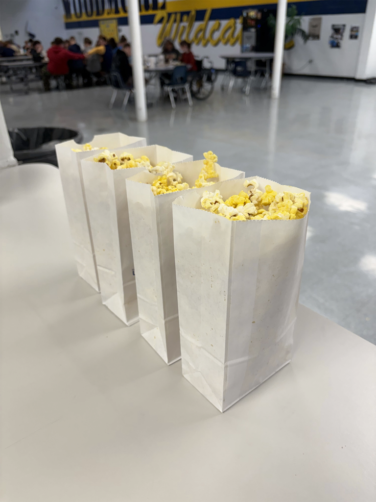 Bags of popcorn wait on a table for students. You can see students eating lunch in the background.