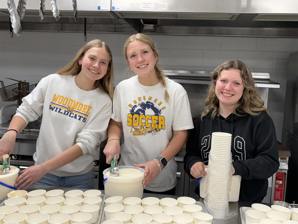 Three students stand at a table in the kitchen, smiling for a picture while scooping ice cream into small bowls.