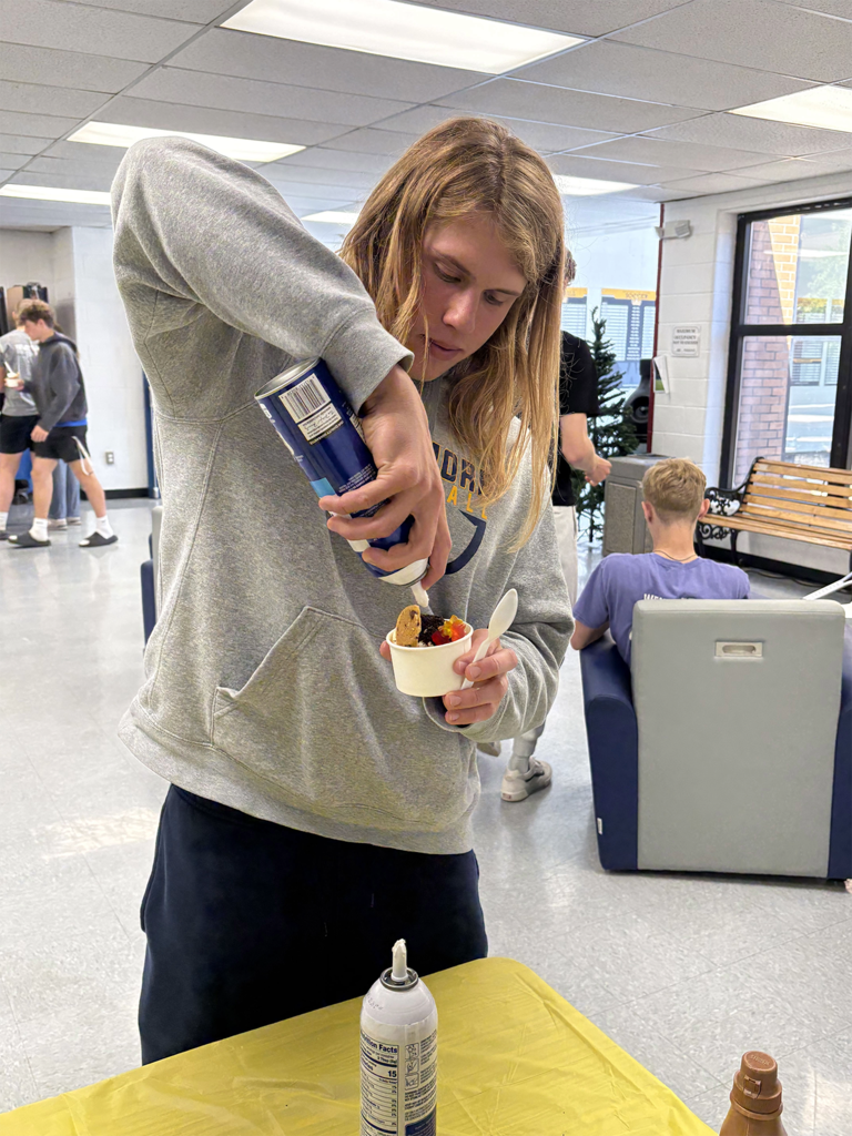 A student is putting whipped cream onto a sundae while other students walk around behind him.