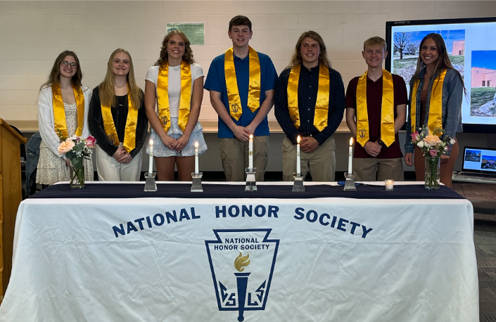 The Senior NHS members stand behind a table with 5 candles and two bouquets of flowers. On the left is a wooden podium with a "W" etched on the front.